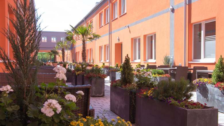 winrooms_gastgarten, © Winrroms Betriebs GmbH A sunny garden with orange buildings, palm trees and colorful flowers in large planters.