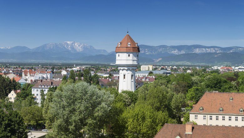 A white water tower with a red roof in a green urban landscape in front of a mountain panorama.