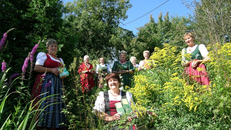 Women in traditional dress stand in a garden with herbs and flowers.