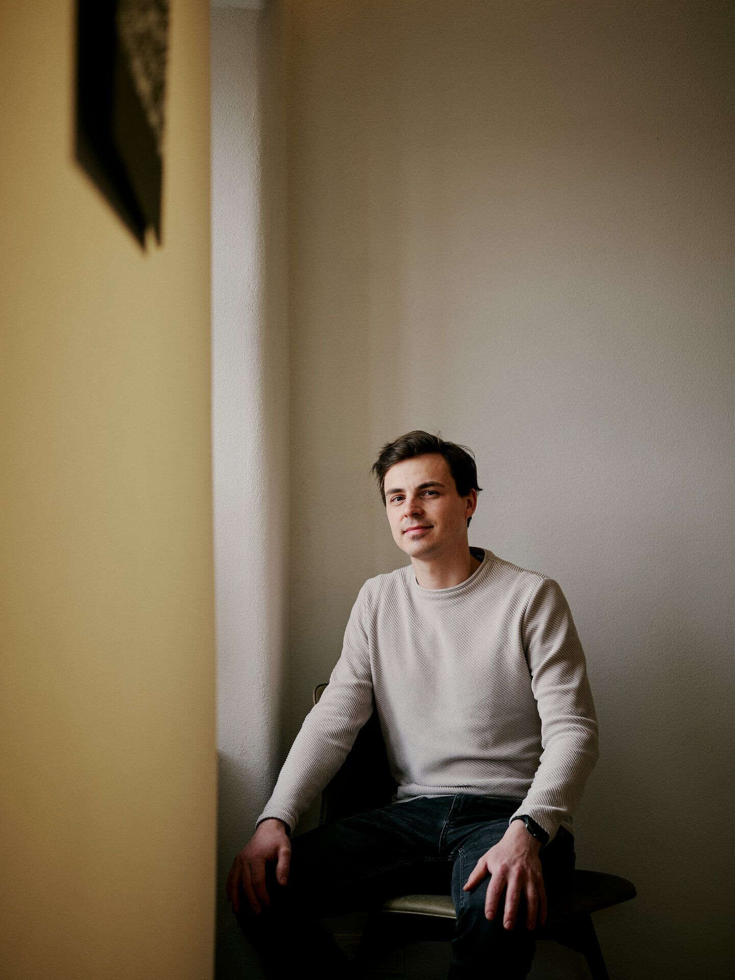 A person sits on a stool in a plain interior and looks into the camera