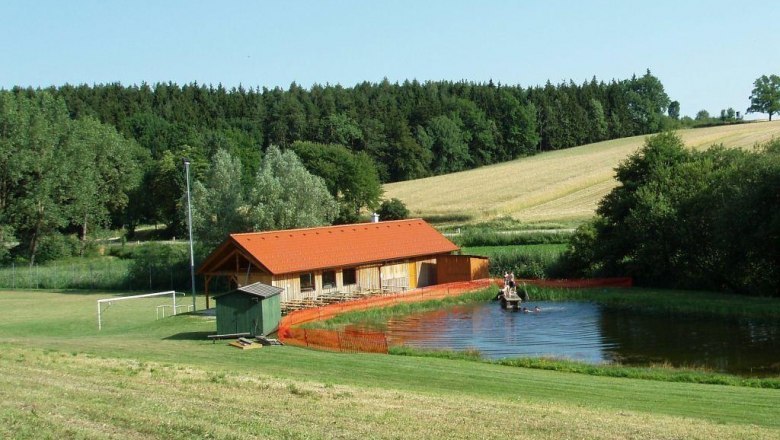 A small bathing pond with a wooden building and surrounding fields and woods.