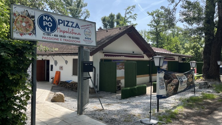 Eingangsbereich aus Sicht des Parkplatzes. Eingangstor mit großem Schild der Pizzeria und Laternen.