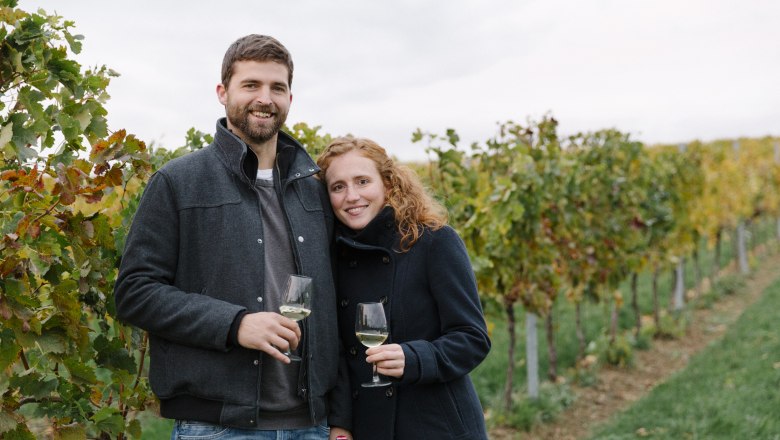 A couple stands in a vineyard holding glasses of wine.