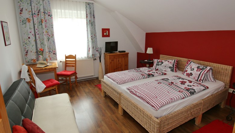 Cozy guest room with double bed, sofa, table and chairs. Red accent wall and floral curtains.