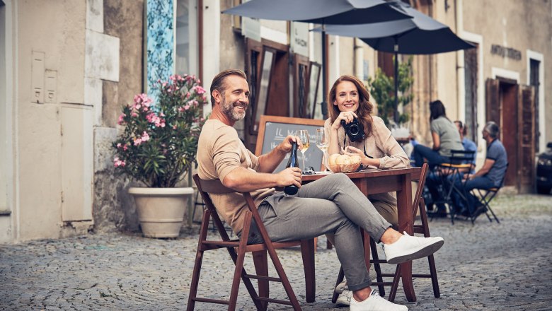 A couple sits at a table in the old town of Krems, drinking wine and smiling. Other guests and historic buildings can be seen in the background.