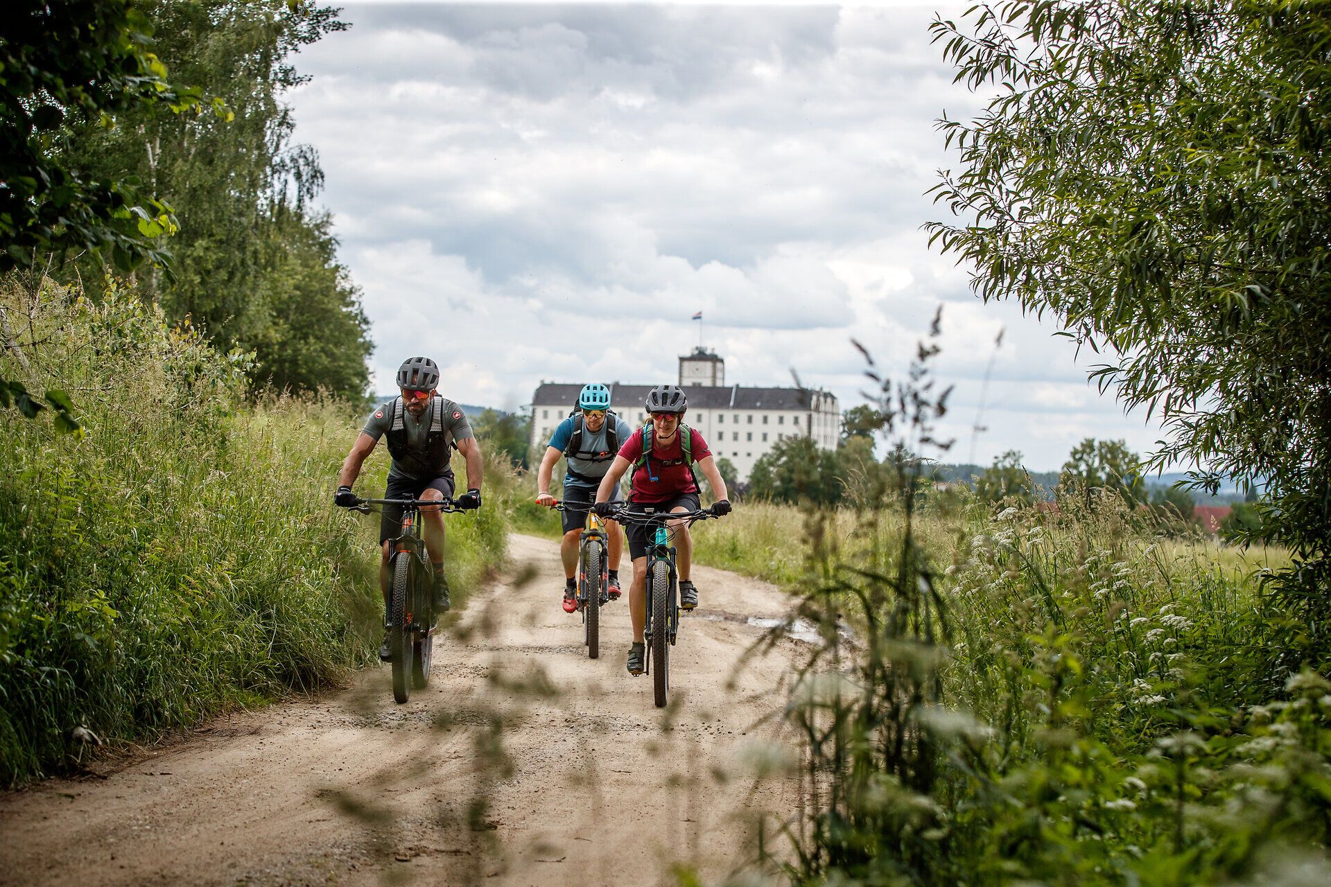 Three mountain bikers glide along a narrow, dusty path surrounded by lush greenery and rolling hills. The fresh mountain air and breathtaking scenery invite you on an unforgettable adventure. Here, where nature and sport meet, every moment becomes an experience.