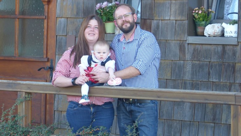 A couple with a baby stands in front of a wooden house.