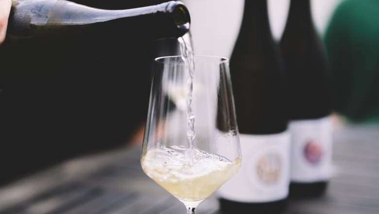 White wine is poured into a glass on a table, with bottles of wine in the background.