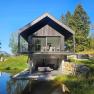 Modern house with wooden facade and balcony, surrounded by nature and a small pond.