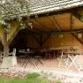 Covered terrace with wooden tables and chairs, surrounded by wooden walls and an apple tree in the foreground.