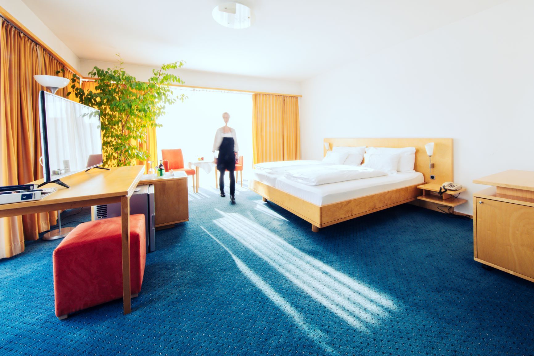 Bright hotel room with double bed, desk, TV and large plant. One person walks through the room.