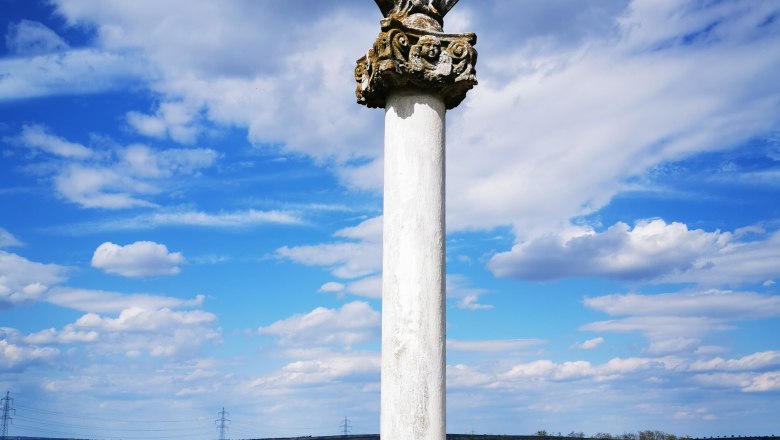 A statue on a pillar in a rural landscape under a blue sky with clouds.