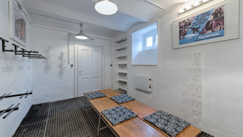 A ski cellar with white walls, ski racks, a wooden bench with cushions and a movie poster on the wall.
