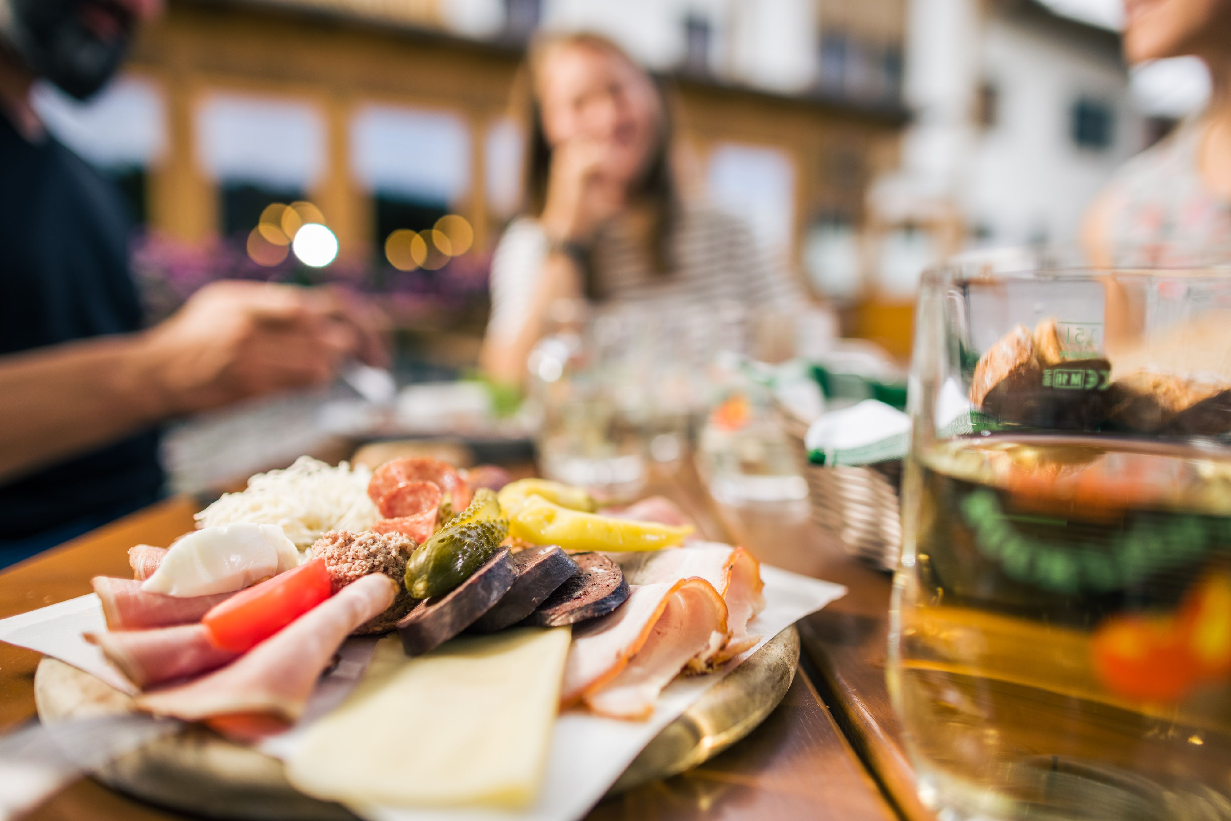 Close-up of a snack with sausage, cheese and vegetables on a table, blurred people in the background.
