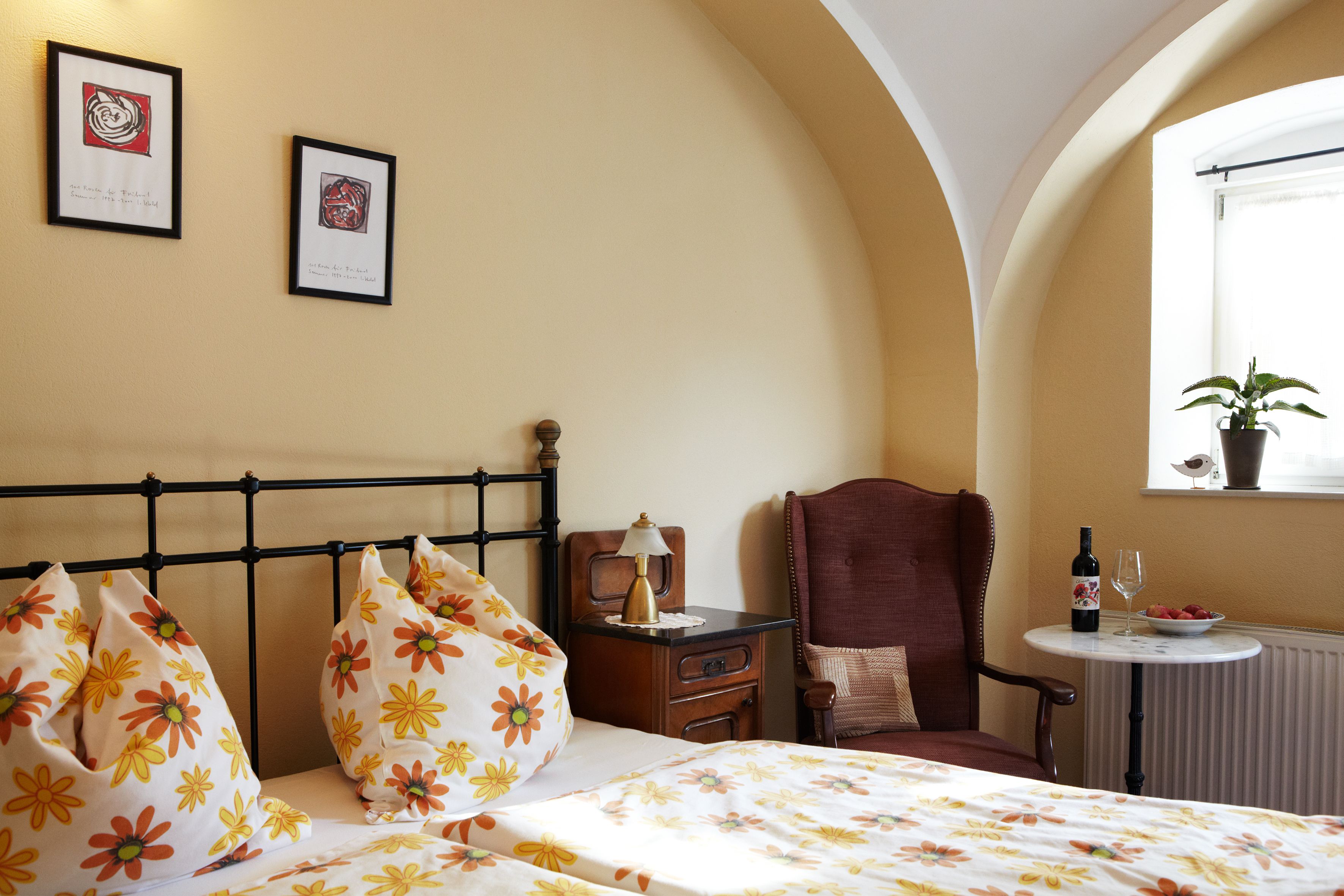 Cozy guest room with floral bedding, armchair, table with wine and fruit, and plants on the windowsill.