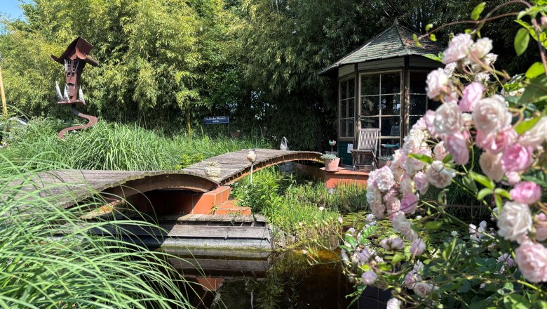 An idyllic garden with a pond, bridge, pavilion and blooming roses in the foreground.