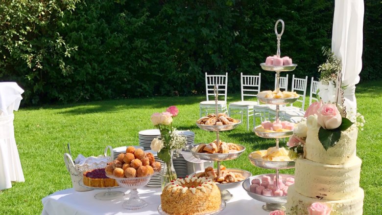 A richly laid table with cakes and pastries in the open air, surrounded by a green lawn and white chairs.