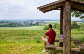 A man sits under a wooden shelter and looks out over a wide landscape with fields and a village in the distance.