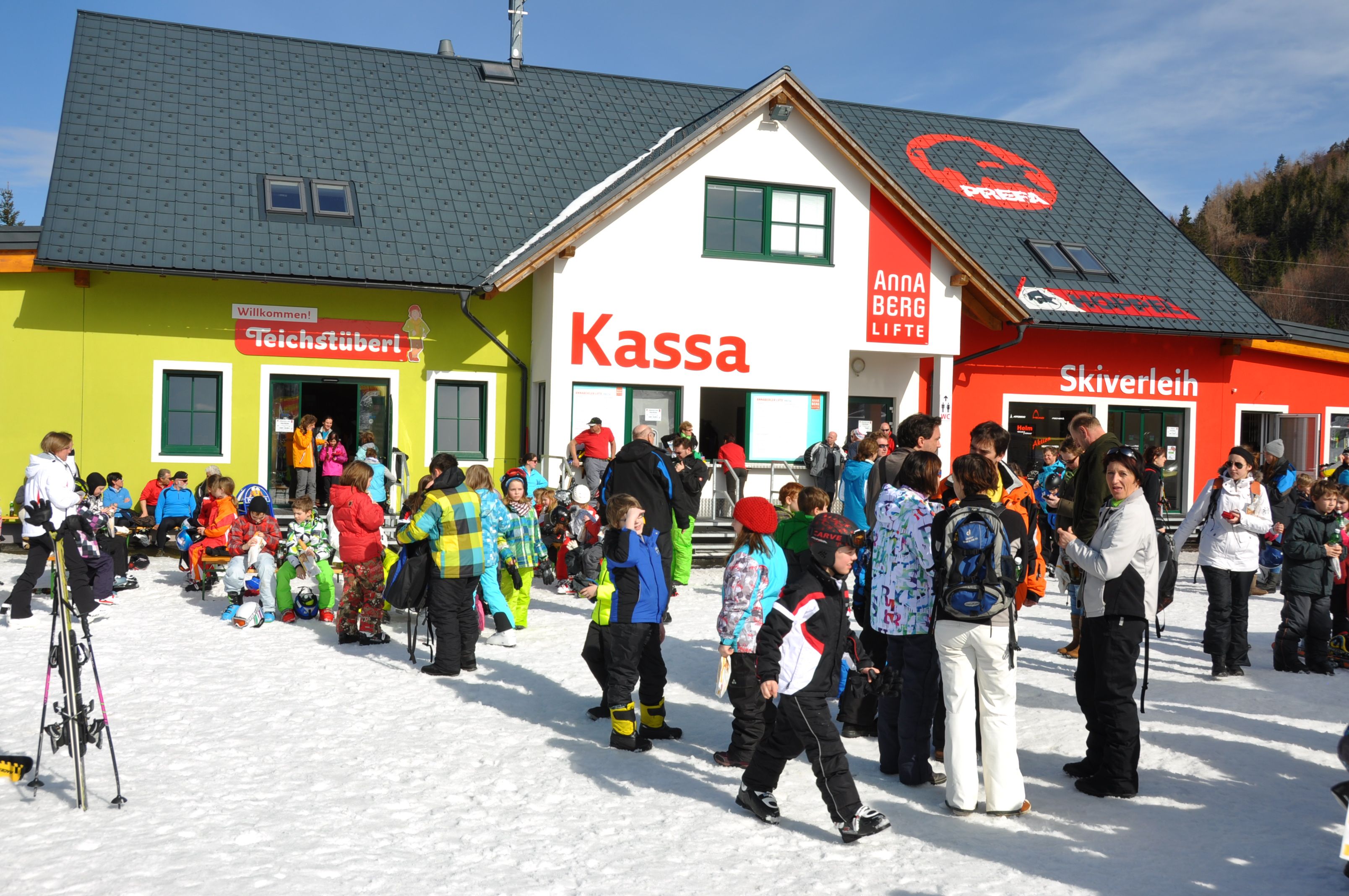 Crowd in front of a ski building with ticket office, ski rental and pond bar.