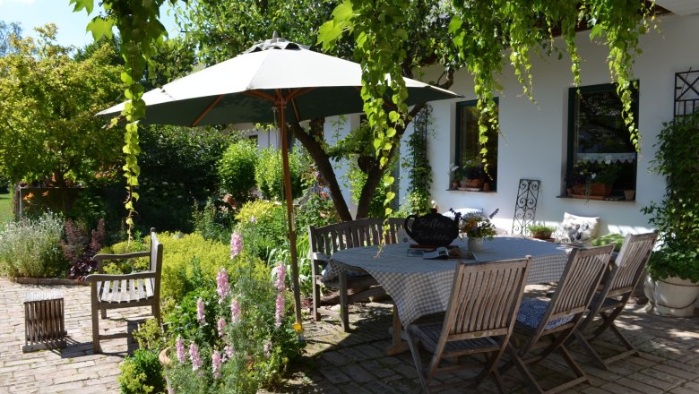 An idyllic garden with a wooden table and chairs under a parasol, surrounded by green plants and flowers.