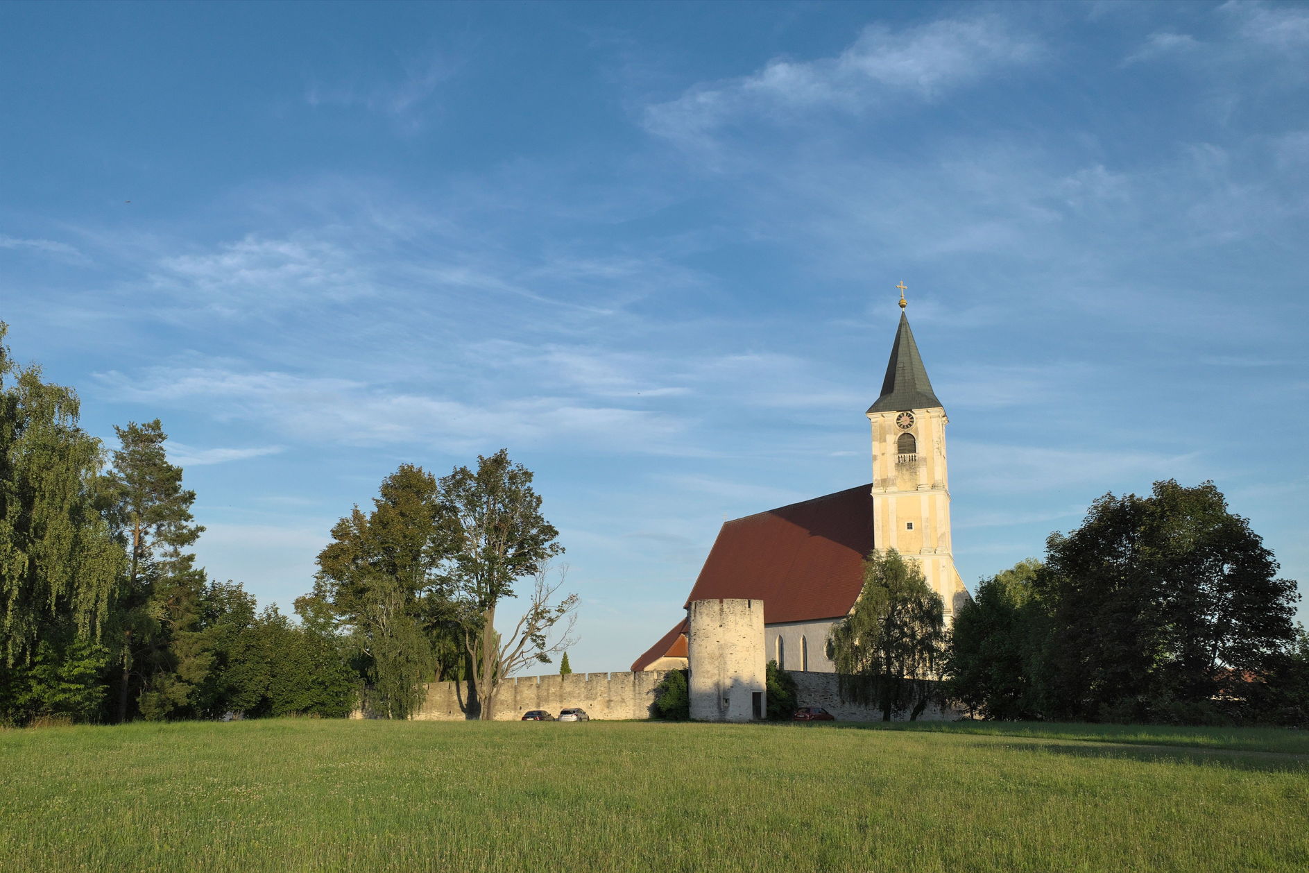 Church with tower and red roof on a meadow, surrounded by trees and a wall.