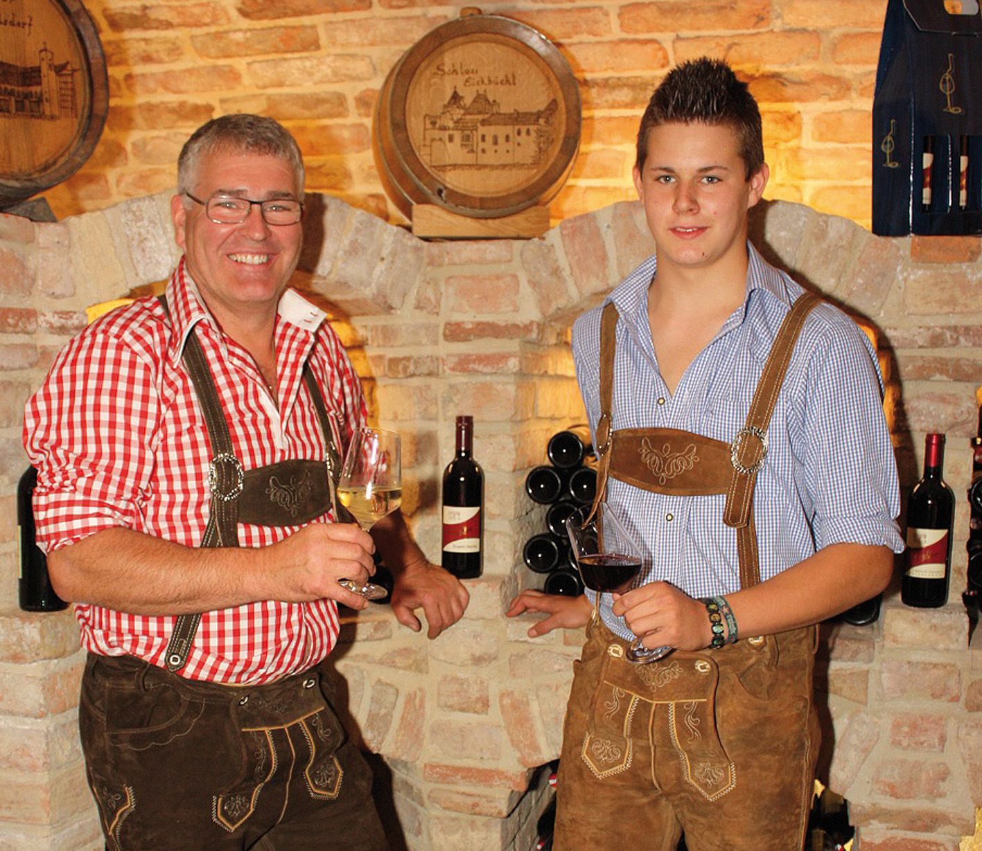 Two men in traditional costume with wine glasses in front of a brick wall.