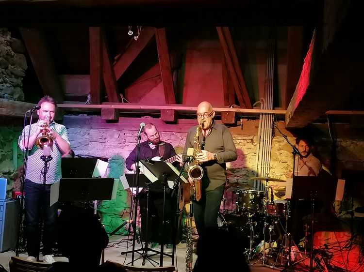 A jazz band plays in a rustic room with wooden beams and stone walls.