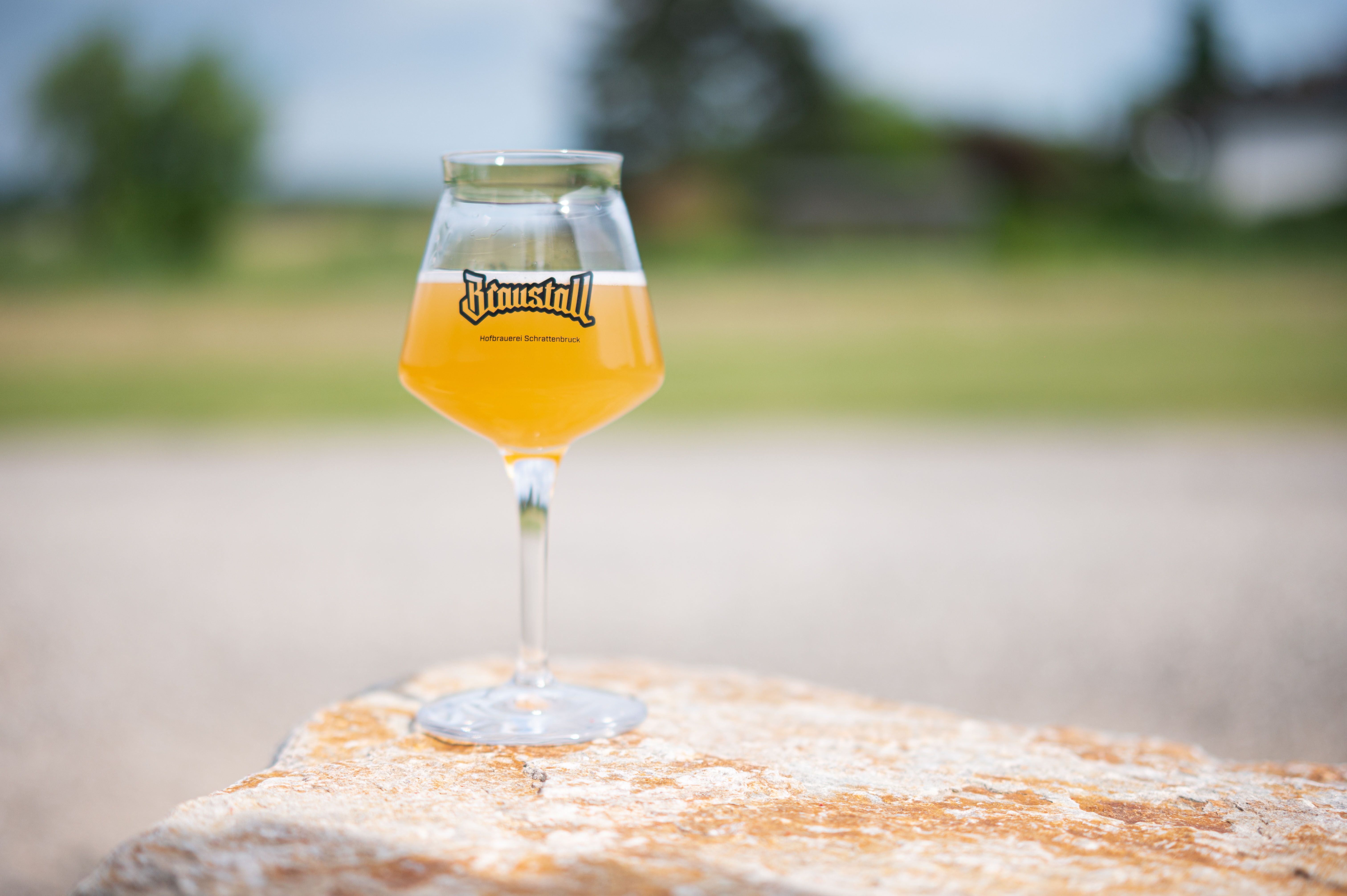A glass of pale beer with the Braustall logo stands on a stone outside.