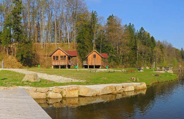 Two wooden huts on the shore of a lake, surrounded by trees and meadows.