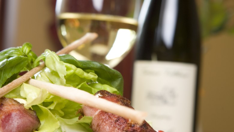 A plate with salad and cheese wrapped in bacon, a wine glass and a bottle of wine in the background.