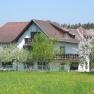 A white house with a brown roof, surrounded by blossoming trees and a green meadow.