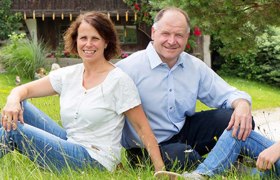 A man and a woman sit smiling on a lawn in front of a house.