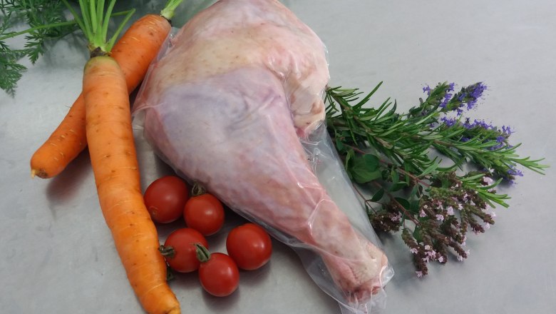 A turkey leg wrapped in plastic with carrots, tomatoes and herbs on a light gray background.