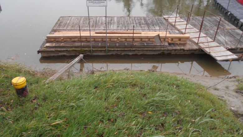 A wooden jetty with a sign and stacked wood, surrounded by water and grass.