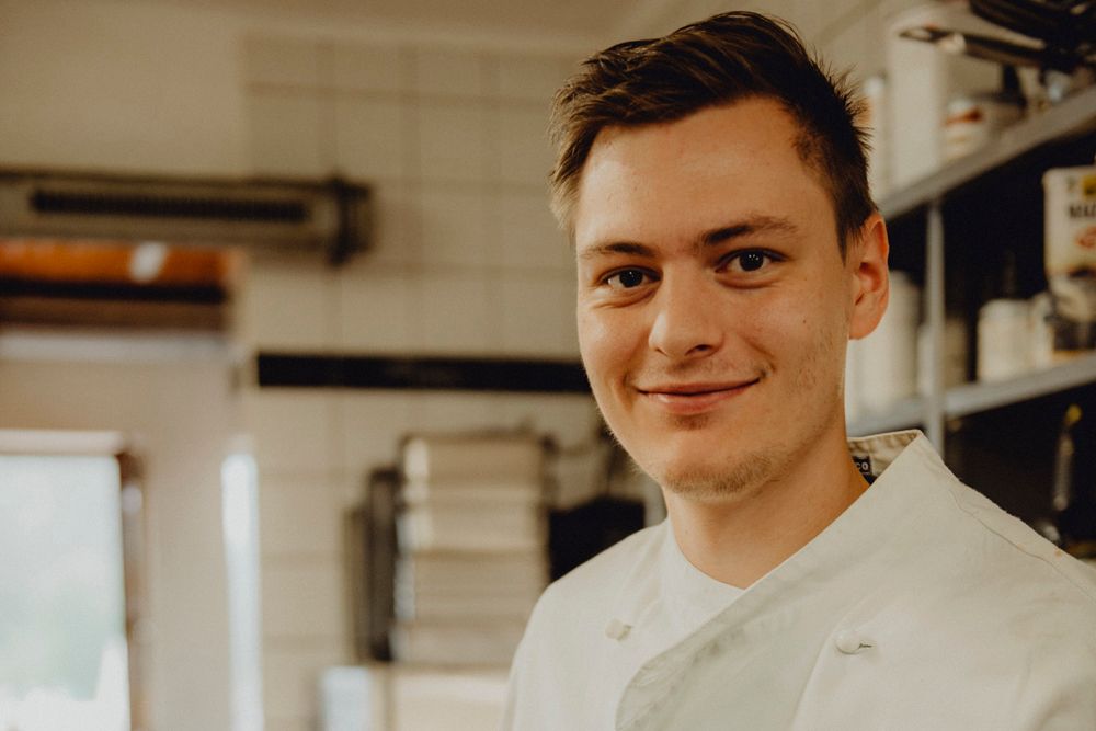 A young man in a white chef's jacket smiles into the camera in a kitchen.