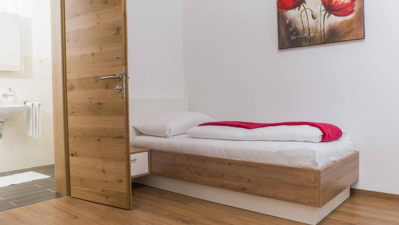 A simply furnished room with a single bed, wooden floor and a picture of red flowers on the wall.