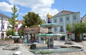 A colorful Hundertwasser-style fountain with golden balls and flowing water, surrounded by buildings and trees.