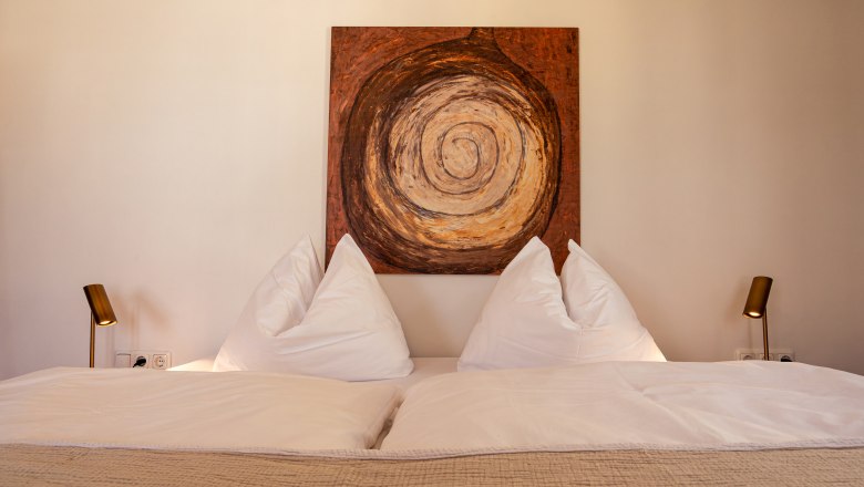 A cozy double bed with white pillows and blankets, flanked by two lamps. An abstract painting with a spiral pattern hangs above the bed.
