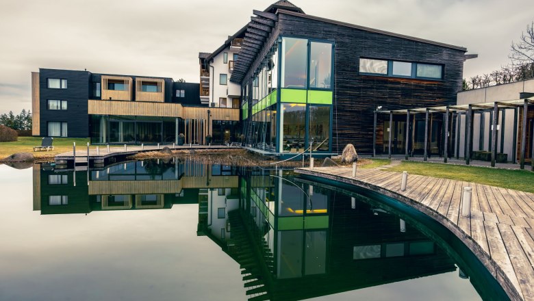 Modern wellness hotel with wooden façade and large pond in the foreground.