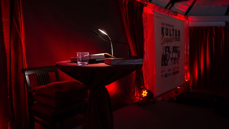 A table with a reading lamp and glass in front of a red curtain, next to it a poster with the inscription 'Kultursommer 2023'.