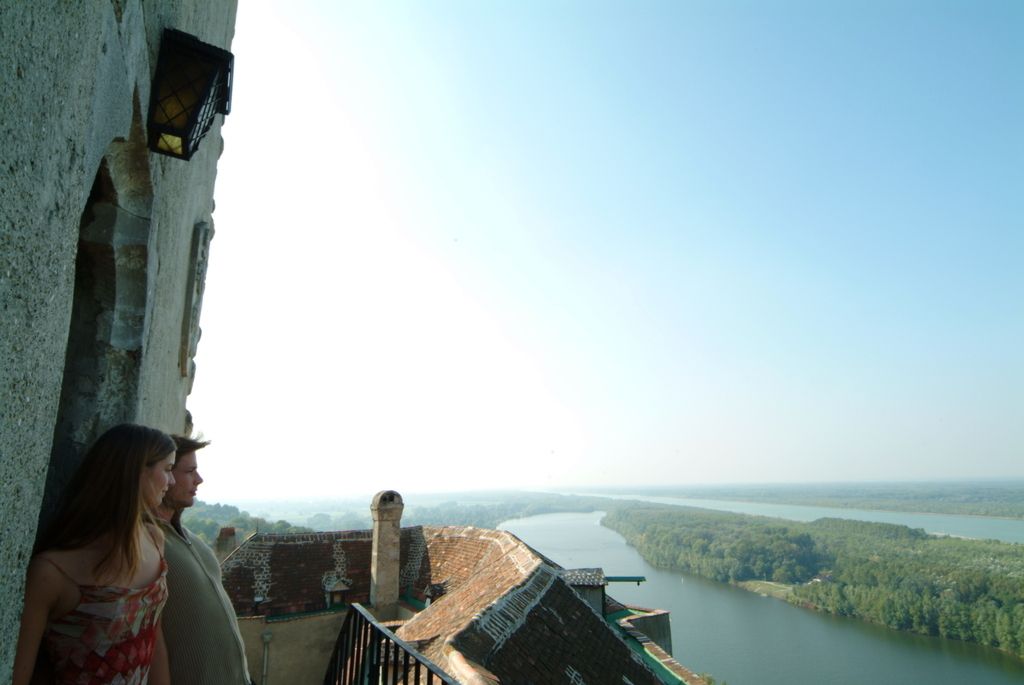 Two people stand on a castle wall and look out over a river and the landscape.