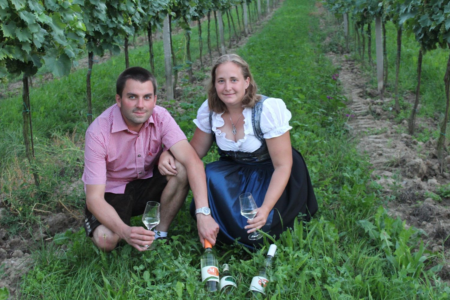 Two people are sitting in a vineyard with wine glasses and bottles.