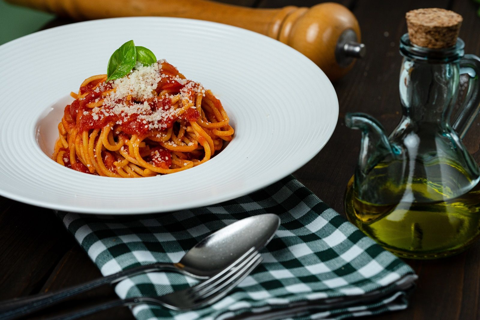 A plate of spaghetti with tomato sauce, basil and parmesan, next to it a bottle of oil and cutlery on a checked cloth.