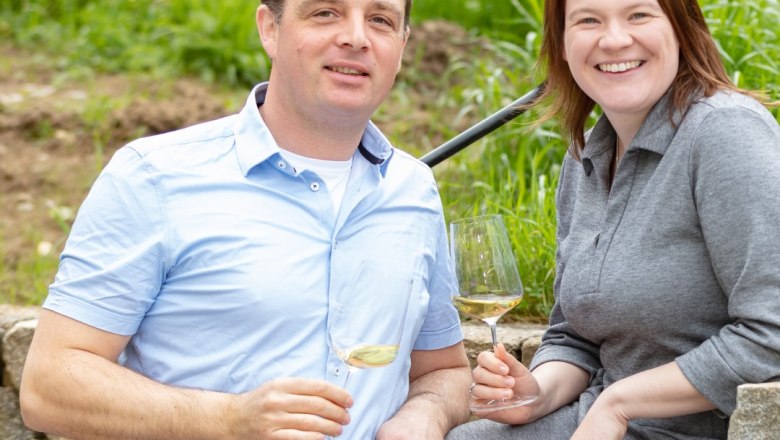 A man and a woman are sitting on a stone wall outside, holding glasses of wine.