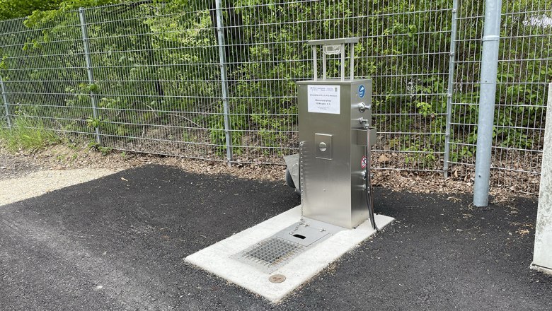 Metal water supply station next to a fence on asphalt.