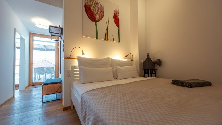 Modern bedroom with double bed, light wooden floor and artwork on the wall. Door leads to a balcony with views of rooftops.