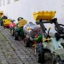 Row of toy tractors on a white wall on cobblestones.