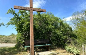 Wooden cross with inscription 'In the cross is salvation' and bench, surrounded by bushes and blue sky.