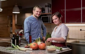 Two people in a kitchen with vegetables and bottles of wine on the worktop.