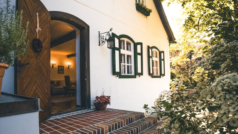 Entrance to a country house with white walls and green shutters, surrounded by flowering plants.
